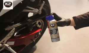 Ręka w czarnej rękawiczce trzyma puszkę smaru do łańcucha motocyklowego K2 przy tylnym kole motocykla.