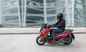 Czerwony skuter z kierowcą w kasku i czarnej kurtce jedzie na tle rozmazanego, nowoczesnego budynku.
