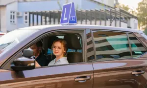 Młoda dziewczyna w okularach za kierownicą auta z literą L, obok instruktor. Pamiętaj, ile ważny jest egzamin teoretyczny na prawo jazdy!