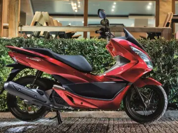 Czerwony skuter Honda PCX, jeden z popularnych **marek skuterów**, zaparkowany przed zielonym żywopłotem.