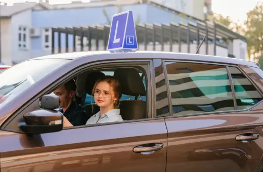 Młoda dziewczyna w okularach za kierownicą auta z literą L, obok instruktor. Pamiętaj, ile ważny jest egzamin teoretyczny na prawo jazdy!
