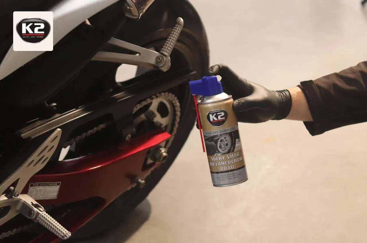 Ręka w czarnej rękawiczce trzyma puszkę smaru do łańcucha motocyklowego K2 przy tylnym kole motocykla.