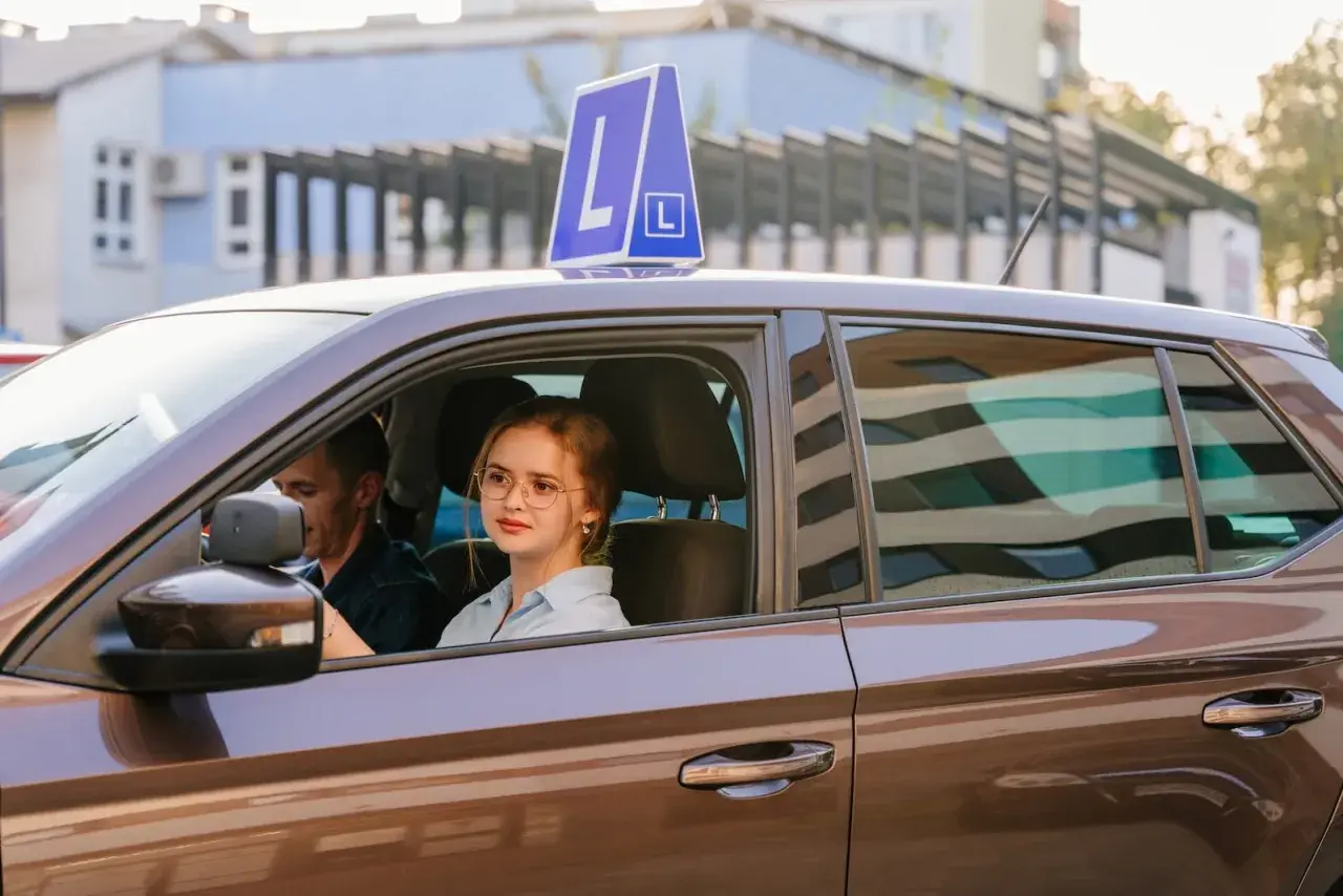 Młoda dziewczyna w okularach za kierownicą auta z literą L, obok instruktor. Pamiętaj, ile ważny jest egzamin teoretyczny na prawo jazdy!