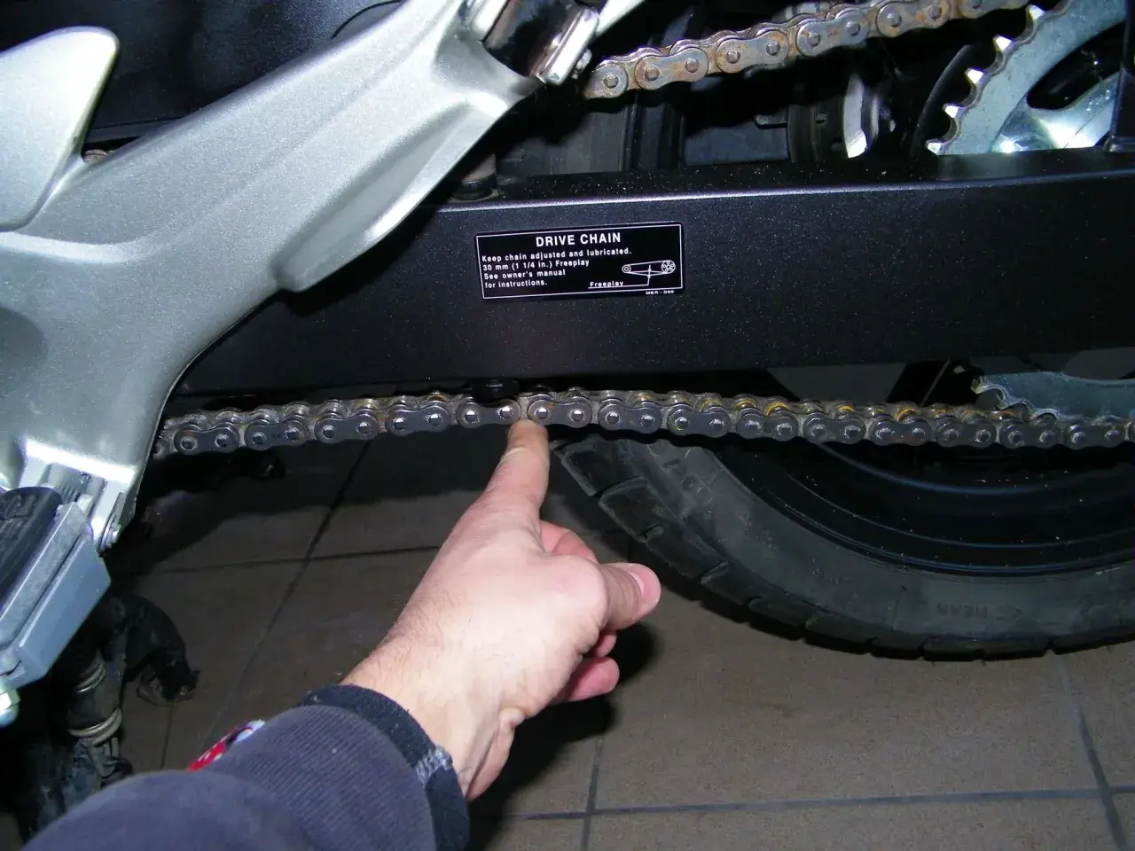 Sprawdzanie luzu łańcucha w motocyklu. Palec pokazuje, jak sprawdzić, czy łańcuch jest prawidłowo naciągnięty.