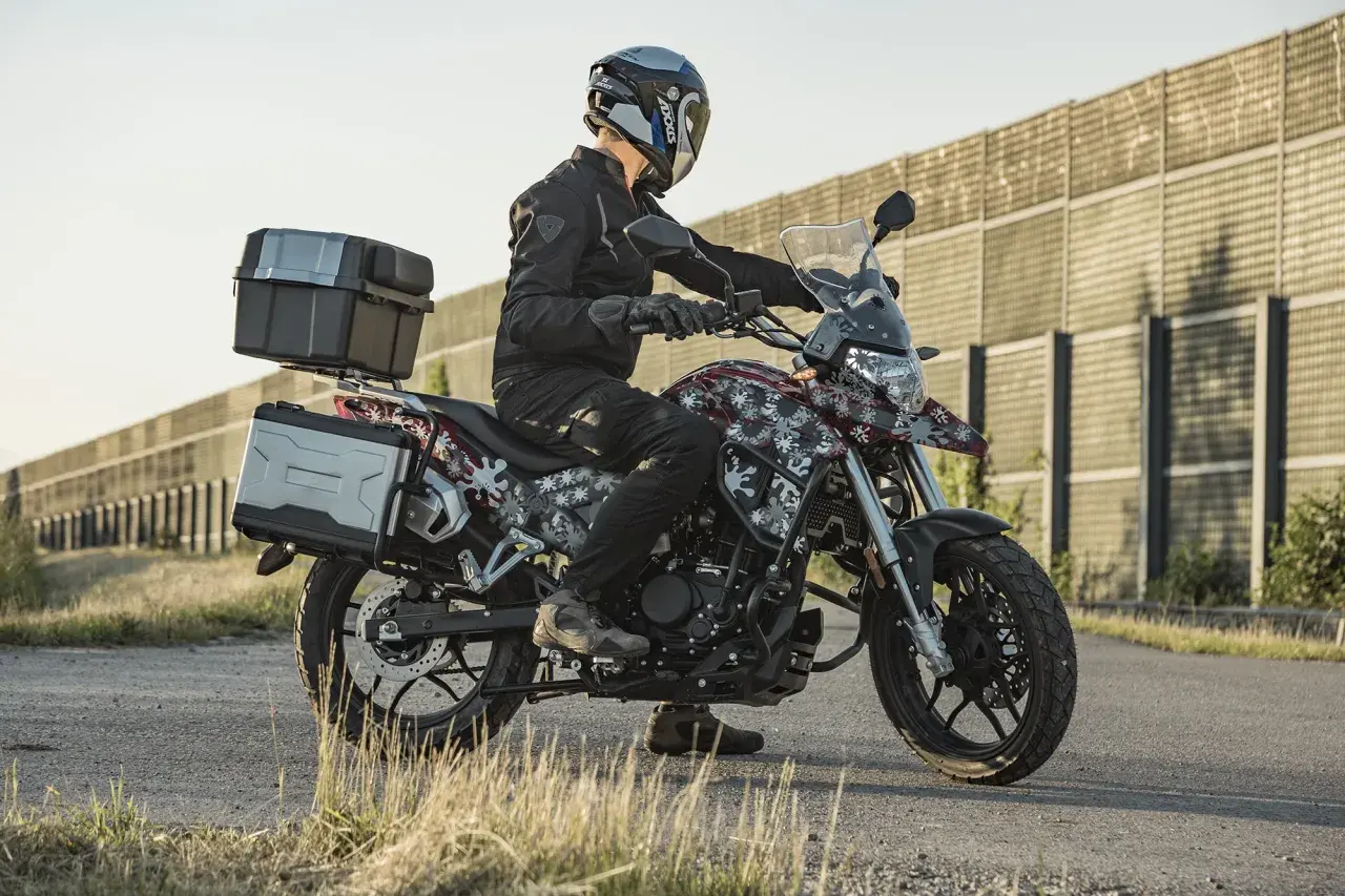 Motocyklista w kasku i czarnej kurtce siedzi na motocyklu Junak RX One 125 z kuframi.
