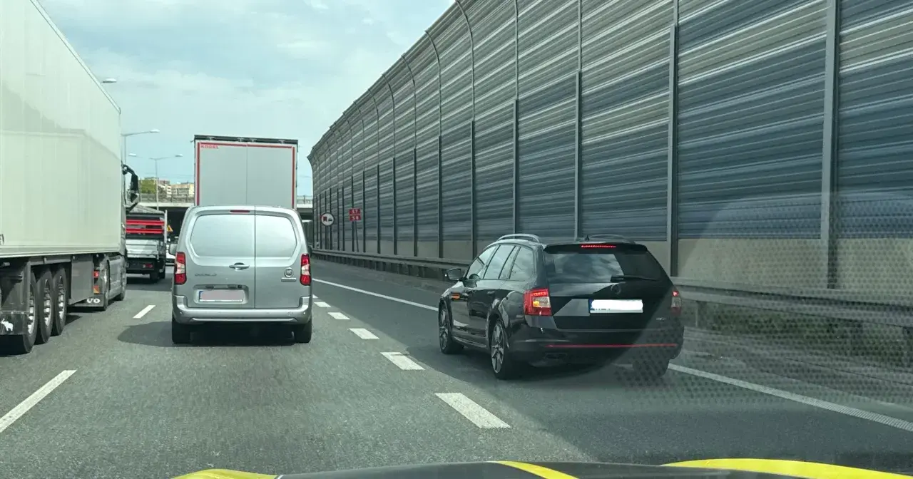 Korek na autostradzie, samochody stoją w rzędzie. Kierowca czarnego kombi zwalnia, by nie dostać mandatu za jazda na suwak.
